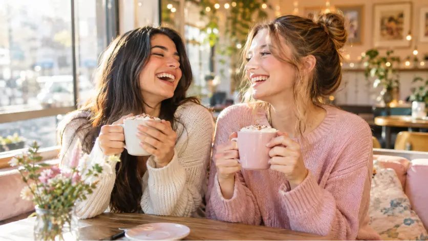 Two best friends laughing together while holding warm drinks in a cozy caf&eacute;